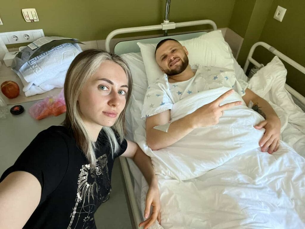 Two people in a hospital room, one resting in a hospital bed and showing a peace sign, symbolizing resilience and recovery.